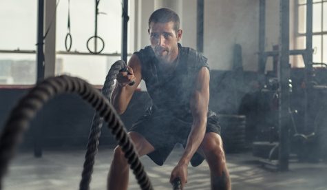 Strong young man working out with battle ropes in a crossfit gym. Muscular sportsman doing cross excursion with ropes in workout gym. Determined guy using battle rope while doing physical training.